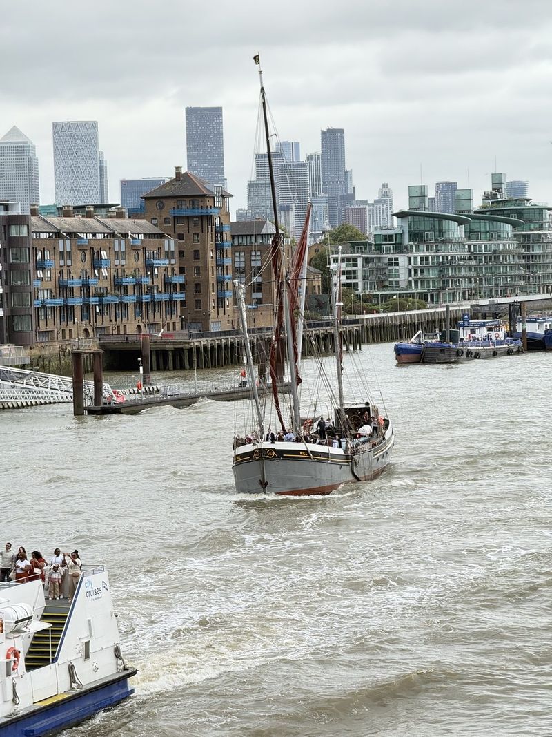 Il veliero tradizionale passa sotto il Tower Bridge aperto, con lo skyline di Canary Wharf sullo sfondo.